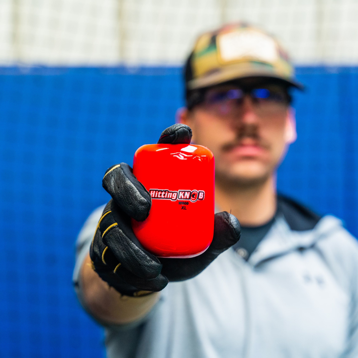 A person in a baseball glove holds the bright red Hitting Knob Bat Weight by Hitting Knob toward the camera. The background is blue and the individual appears slightly out of focus.