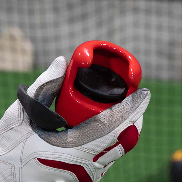 A person wearing white and red sports gloves holds The Hitting Knob Bat Weight by Hitting Knob with a black cap, in front of a blurred sports net and green turf background.