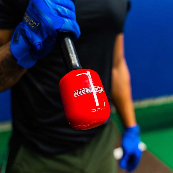 A person in blue gloves holds a black bat fitted with the Hitting Knob Bat Weight by Hitting Knob, a baseball hitting aid designed to improve bat speed, against a blurred indoor sports background.