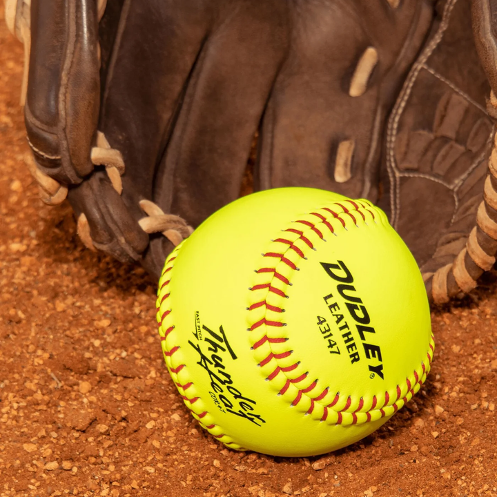 A Dudley Thunder Heat 11" 47/375 Leather Fastpitch Softball (43531N) by Dudley rests on brown dirt beside a worn glove, ready for the next game.