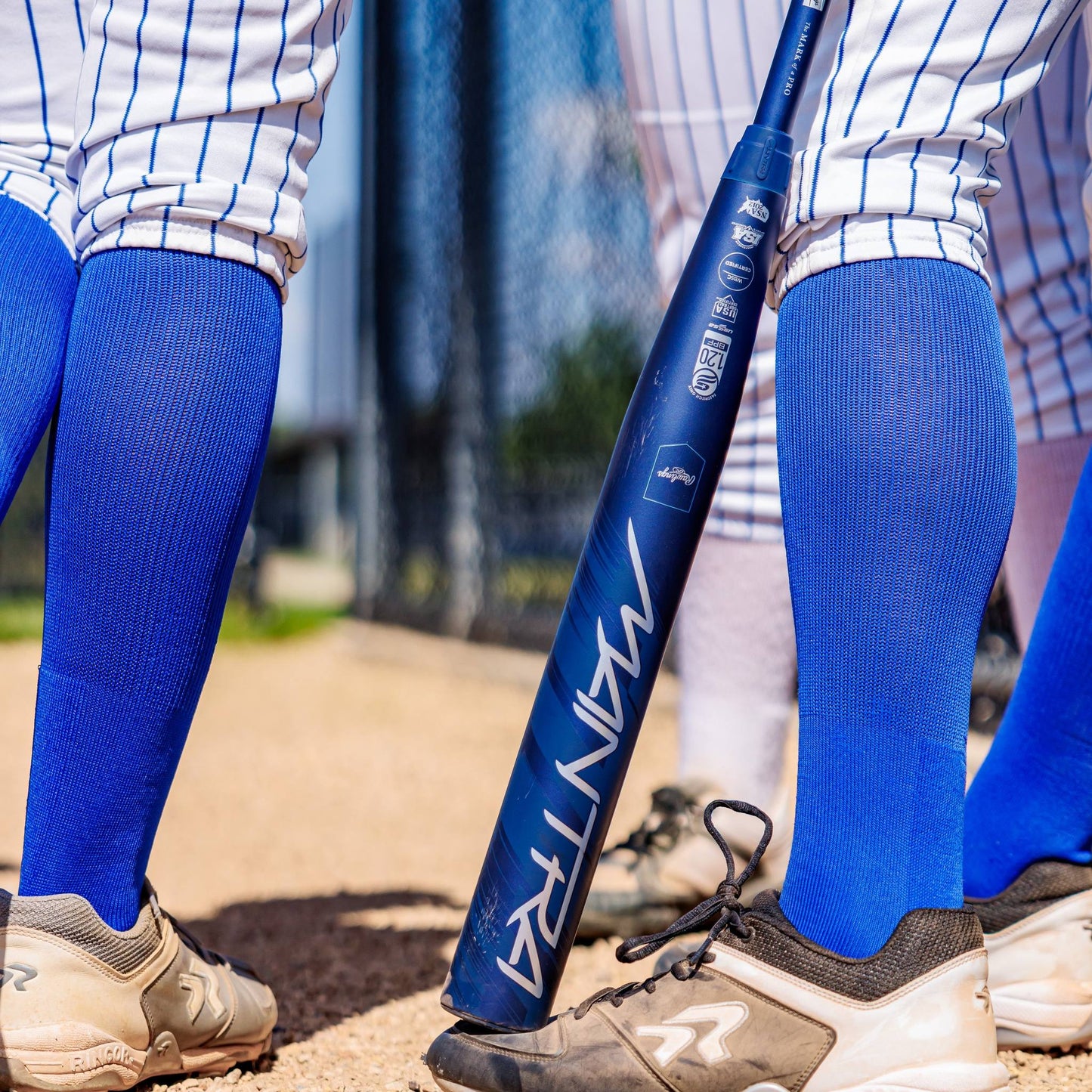 Several athletes in blue knee-high socks, white pinstripe pants, and cleats stand on a dirt field. A 2025 Rawlings Mantra (-10) Fastpitch Softball Bat: RFP4M10 (DEMO) from Rawlings leans against one player’s leg.