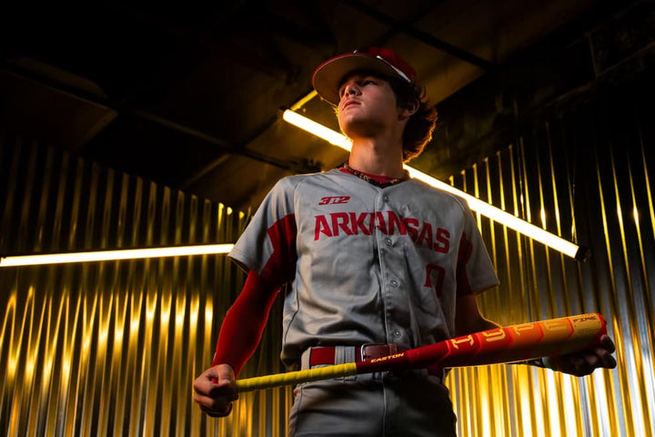 An Arkansas player grips the 2025 Easton Hype Fire (-8) 2 5/8" USA Baseball Bat (EUS5HYP8) by Easton, looking upward as bright yellow lights reflect off metallic walls for a dramatic, intense scene.