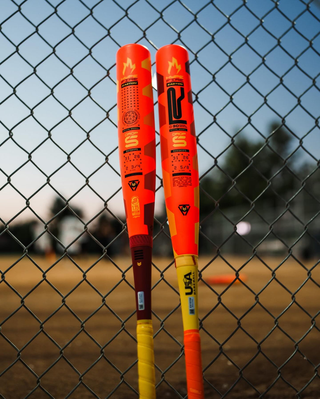 Two vibrant Easton 2025 Hype Fire (-8) 2 5/8" USA Baseball Bats (EUS5HYP8) lean against a chain-link fence, with a blurred baseball field in the background, showcasing bold graphics perfect for power-hitting young players.
