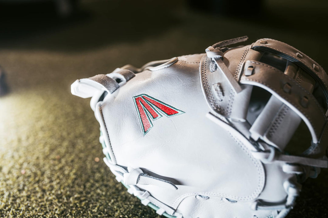 A close-up of the Easton Jen Schro "The Rudi" 34" Fastpitch Catcher's Mitt (RUDI-2024) made from premium USA steerhide, with a red and black stitched logo, rests on a textured green surface under bright lighting.