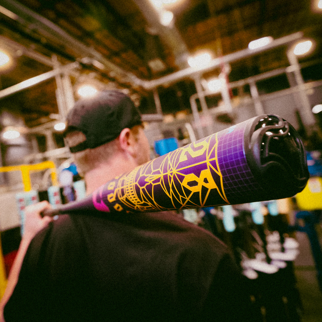 A person in a black cap holds a colorful, purple and yellow baseball bat over their shoulder in an indoor, industrial-looking setting. The focus is on the detailed design of the bat.
