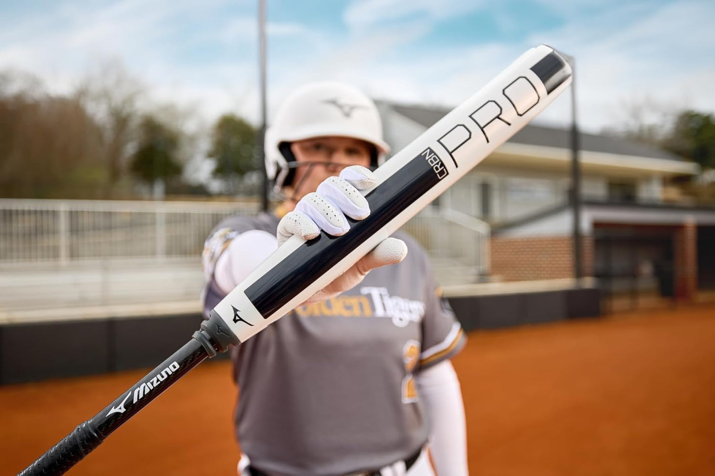 A softball player in uniform and helmet stands on a dirt field, holding the 2025 Mizuno CRBN Pro (-10) Fastpitch Softball Bat (340665) toward the camera, with a slightly blurred background.