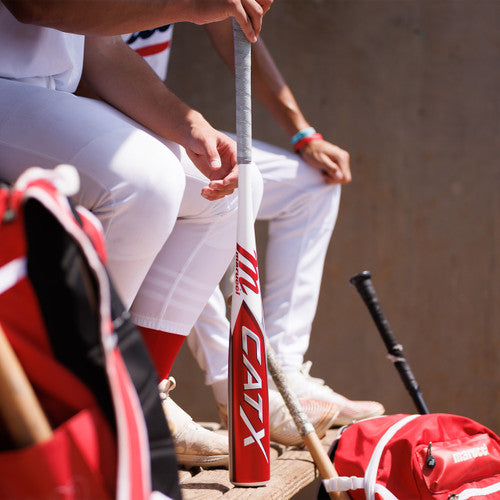 Two baseball players in white uniforms sit on a bench, one holding a red and white CATX bat vertically. Baseball gear, including bats and a red bag, are nearby.