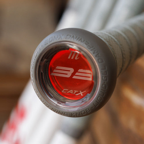 Close-up of the end cap of a baseball bat, featuring a red circular design with “33” and “CATX” text visible, surrounded by gray text on the knob, with other bats blurred in the background.