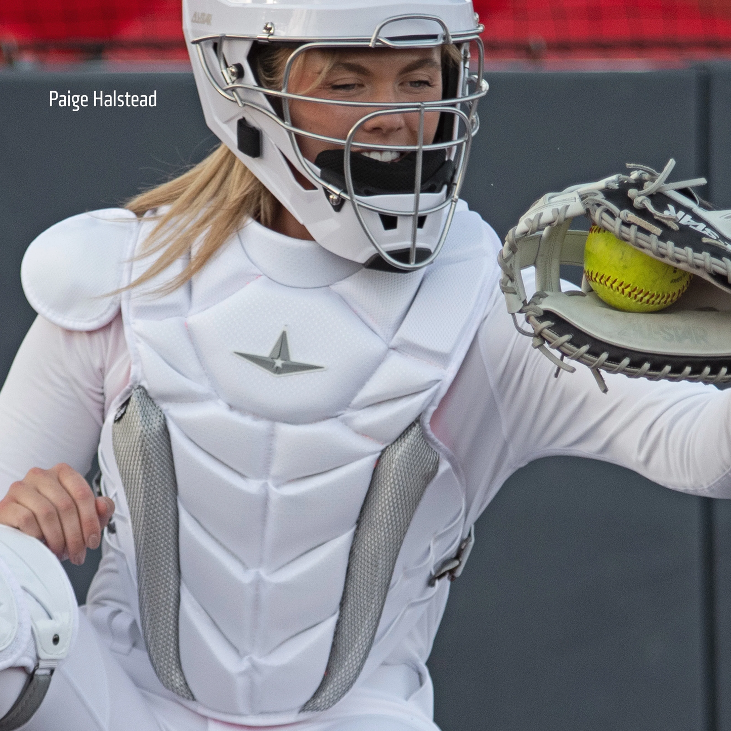 A woman in a white uniform holds a softball, wearing the All Star PHX Paige Halstead Fastpitch Catcher's Chest Protector (CPW-PHX) by All Star.