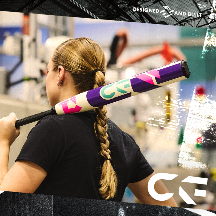 A person with a long braided ponytail holds the 2026 DeMarini CF (-11) Fastpitch Softball Bat: WBD2630010 over their shoulder indoors. The colorful purple bat features pink, green, and white designs, plus a Paraflex Plus Composite Barrel.