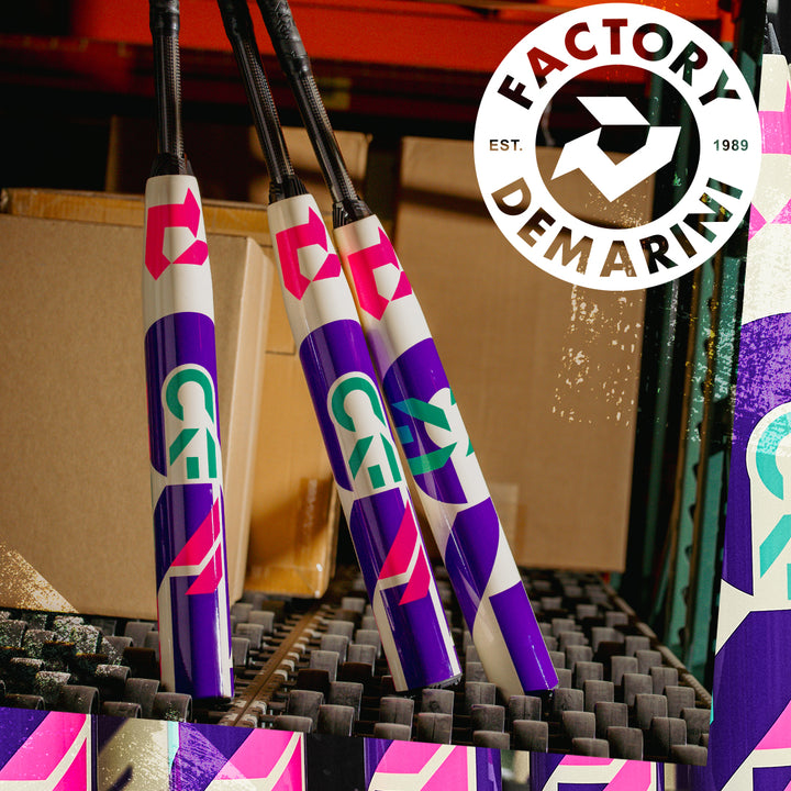 Three vibrant 2026 DeMarini CF (-11) Fastpitch Softball Bats with purple, white, pink, and green designs stand upright against stacked boxes in a warehouse. Paraflex Plus Composite Barrels and the DeMarini logo are also visible.
