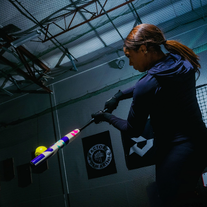 A woman in athletic wear swings a 2026 DeMarini CF (-11) Fastpitch Softball Bat (WBD2630010) at a yellow softball inside an indoor training facility with netting and banners on the walls.
