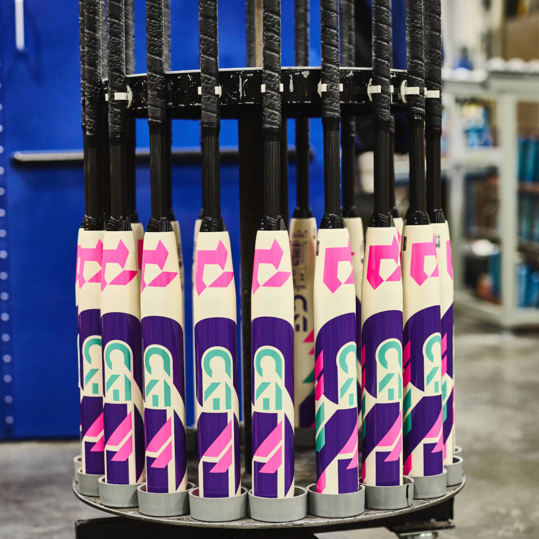 A rack displays multiple colorful DeMarini bats, including the 2026 DeMarini CF (-11) Fastpitch Softball Bat: WBD2630010, with Paraflex Plus Composite Barrel and vibrant geometric designs in purple, pink, teal, and white, all standing vertically.