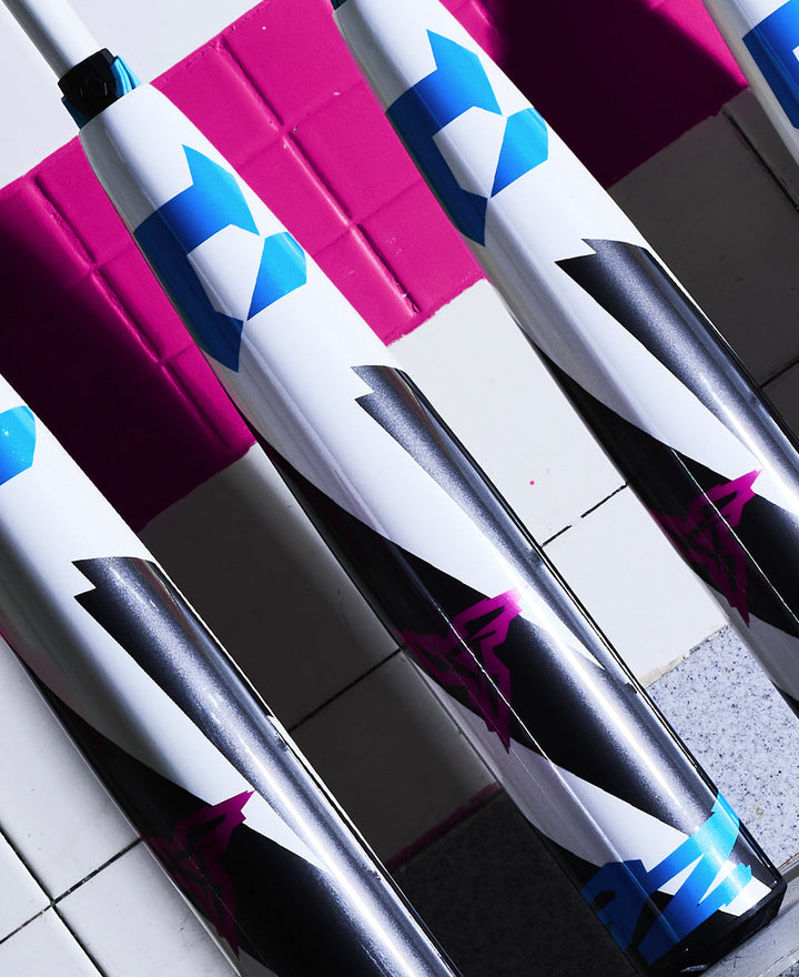 Close-up of several used 2025 DeMarini ZEN (-10) 2 3/4" USSSA baseball bats (WBD2532010) with blue, black, and magenta geometric designs by DeMarini, arranged diagonally on a pink and white tiled surface with bold colors and sharp shadows.
