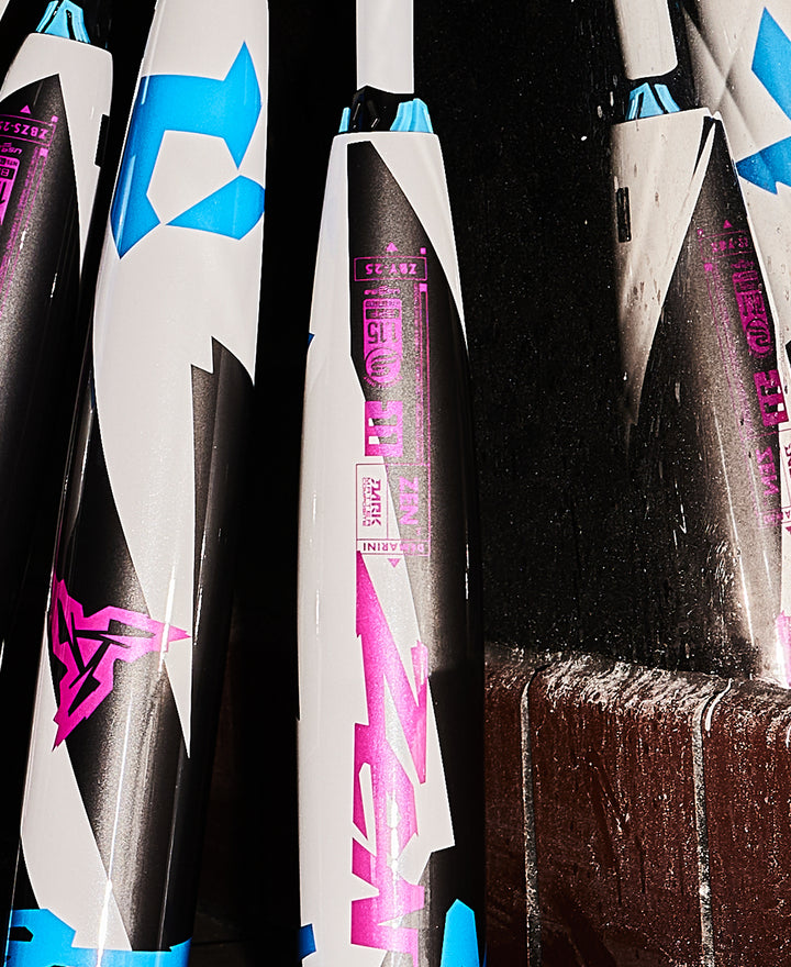 Close-up of several DeMarini USSSA Baseball Bats, including the 2025 DeMarini ZEN (-5) 2 3/4" (WBD2534010), with bold white, black, blue, and pink graphics propped against a dark wall and chalk residue on the surface below.