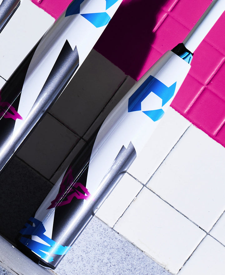 Two white 2025 DeMarini ZEN (-8) USSSA Baseball Bats, featuring blue, black, and pink geometric designs, stand upright on a tiled floor with white and pink tiles, highlighting their sleek Dark Matter Composite construction.