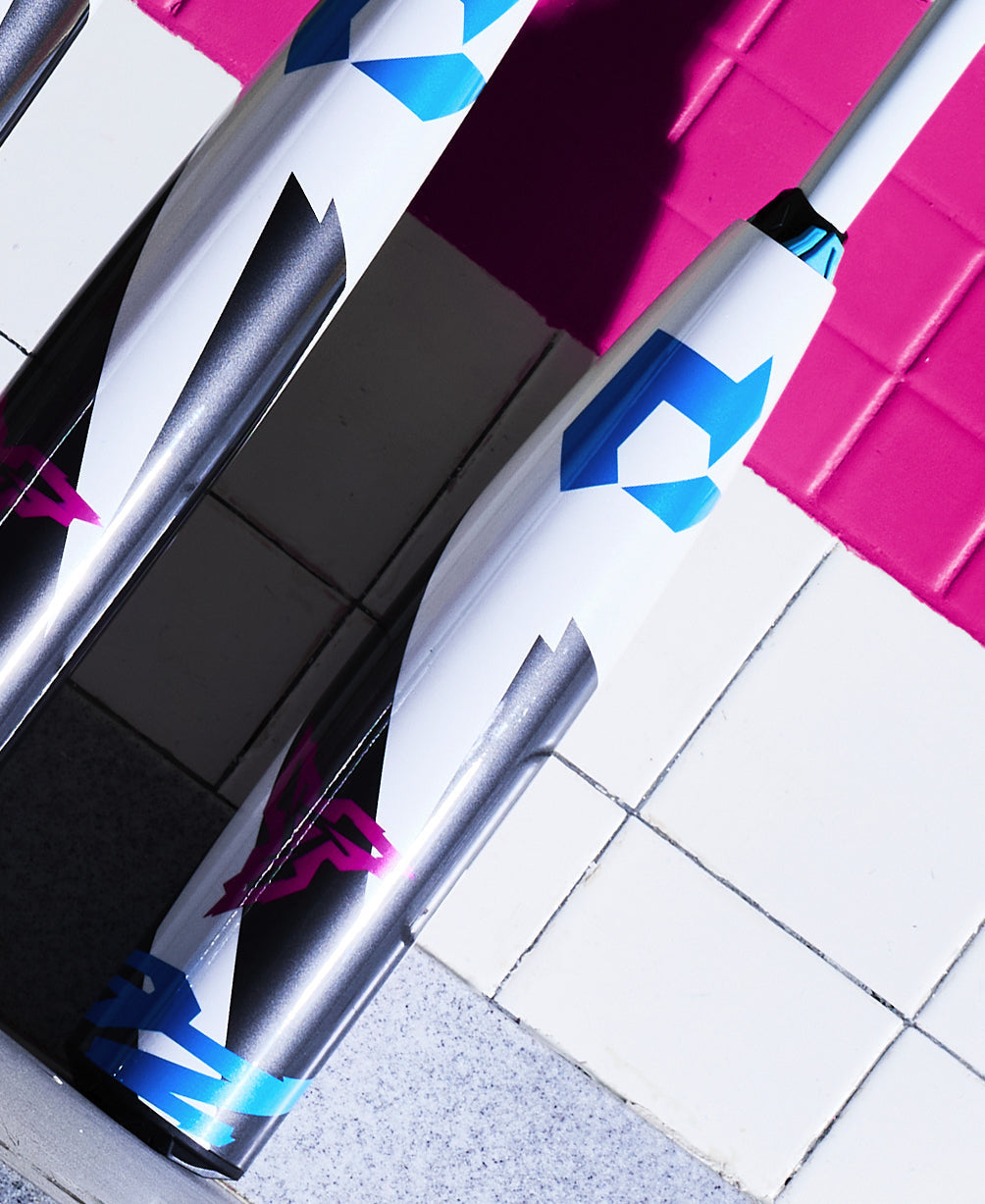 Two modern USSSA baseball bats, including the 2025 DeMarini ZEN (-10) with Dark Matter Composite and geometric blue, black, and pink designs, lean against a white and pink tiled wall.