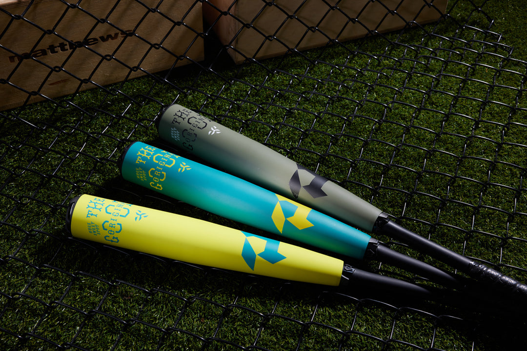 Three colorful bats—gray, teal, and yellow—rest on artificial grass near a chain-link fence, including the 2025 DeMarini The Goods Teal (-3) BBCOR Baseball Bat (WBD2564010), with wooden crates in the background.