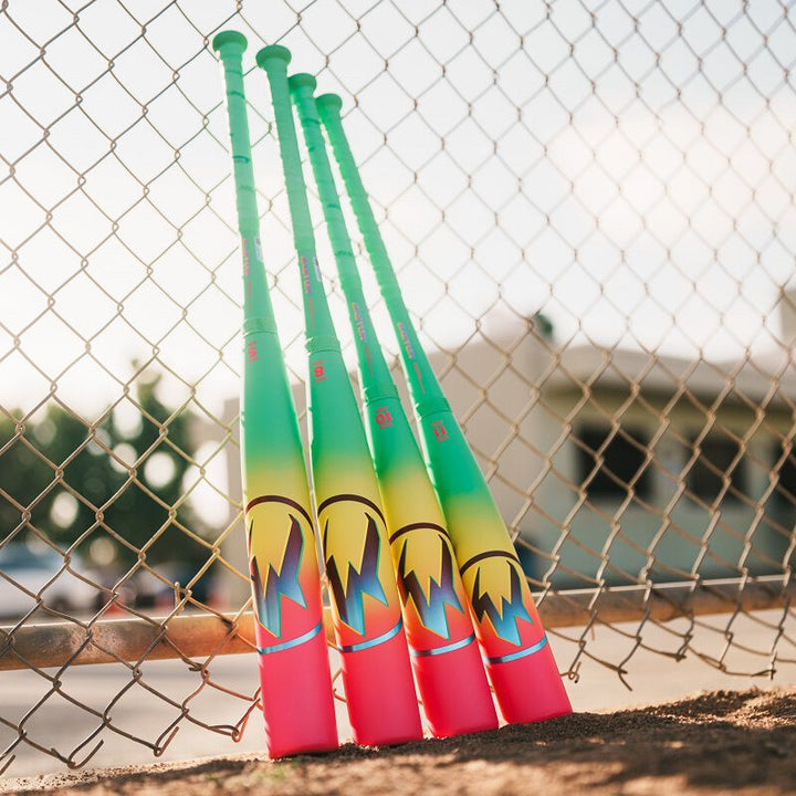 Four neon baseball bats, including the 2026 Easton Hype Fire (-11) USA Baseball Bat by Easton with a bright green handle, lean against a chain-link fence on a sunny day, with blurred buildings and trees in the background.