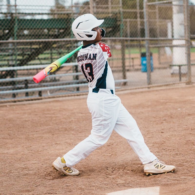 A young baseball player in a white uniform and helmet swings the 2026 Easton Hype Fire (-11) 2 5/8" USA Baseball Bat (EUS6HYP11) on a dirt field, with a chain-link fence and empty bleachers in the background.