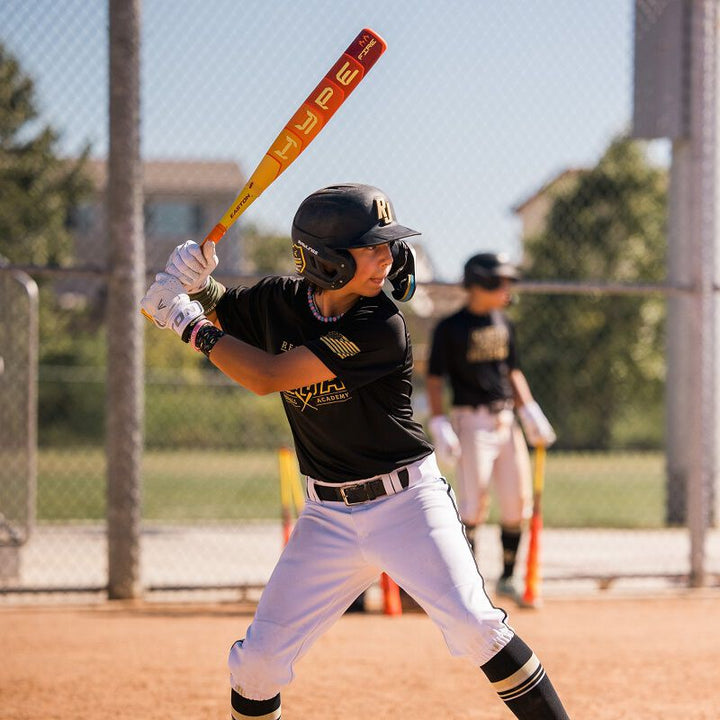 A young ballplayer in a black helmet grips his 2025 Easton Hype Fire JBB (-10) 2 3/4" USSSA Baseball Bat (EJB5HYP10) at home plate on a sunny field, with another player visible in the background.