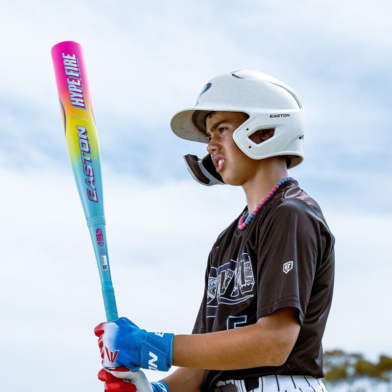 A young baseball player in a helmet and gloves grips an Easton 2026 Hype Fire (-5) 2 3/4" USSSA Baseball Bat (EUT6HYP5), looking focused in his dark jersey with white lettering beneath a bright, cloudy sky.