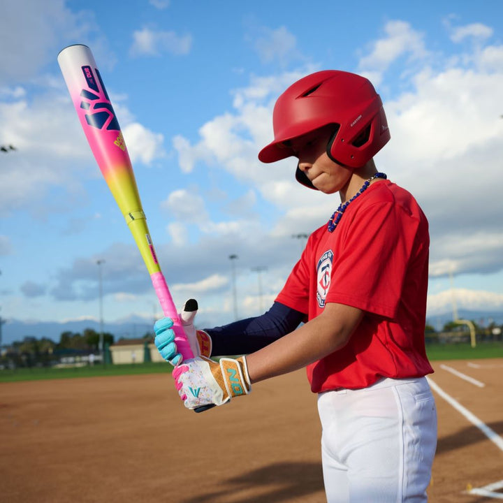 A young player in a red helmet stands on the field gripping the Easton 2026 Dub (-10) USSSA Baseball Bat (EUT6DUB10), a vibrant two-piece composite bat, ready to swing under a blue sky dotted with clouds.