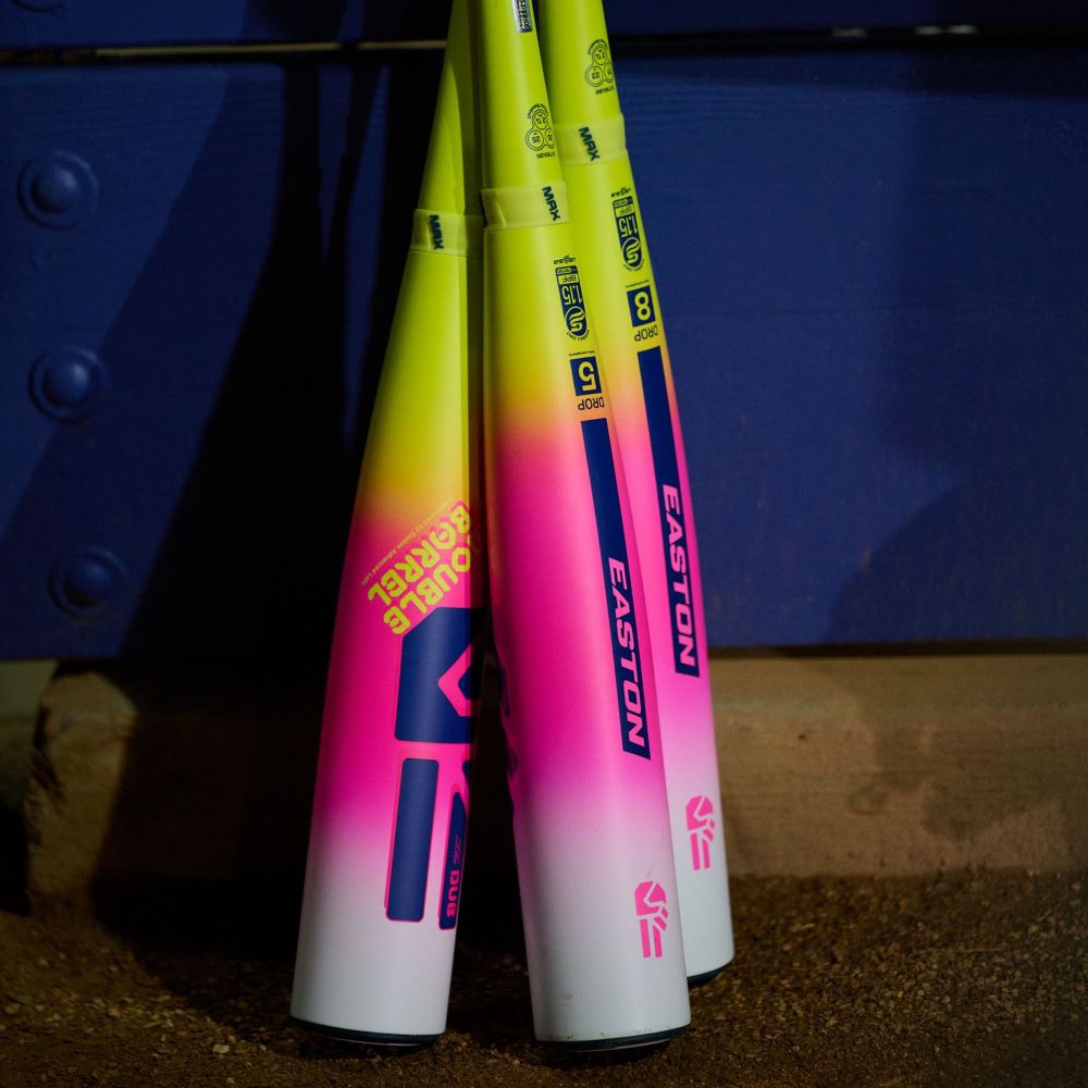 Three 2026 Easton Dub (-10) 2 3/4" USSSA Baseball Bats, featuring a yellow-to-pink gradient and Double Barrel two-piece composite design, lean against a blue wall on dirt. DOUBLE BARREL text is visible on one bat.