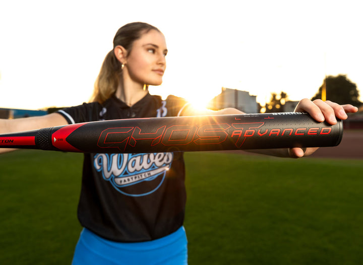 A softball player in a Waves jersey holds out the 2024 Easton Ghost Advanced (-9) Fastpitch Softball Bat (EFP4GHAD9), its black and red design shining as the sun sets over a grassy field.