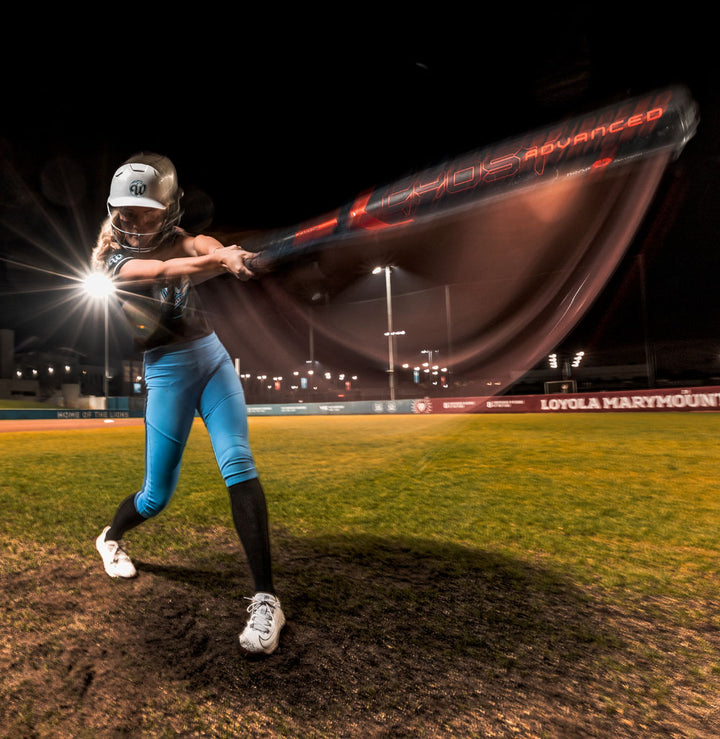 A softball player in blue pants and a helmet swings the 2024 Easton Ghost Advanced (-9) Fastpitch Softball Bat by Easton at night on a lit field, with motion blur showing speed and "Loyola Marymount" visible on a sign in the background.