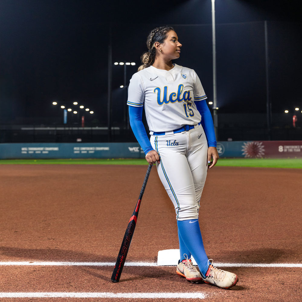 A UCLA softball player in full uniform stands on a field at night, leaning on the Easton 2024 Ghost Advanced (-9) Fastpitch Softball Bat (EFP4GHAD9), illuminated by stadium lights.