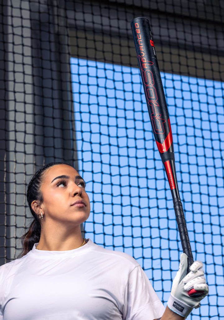 A woman in a white shirt and batting glove holds the 2024 Easton Ghost Advanced (-9) Fastpitch Softball Bat (EFP4GHAD9) upright, gazing at it with a netted fence and blue sky behind her.