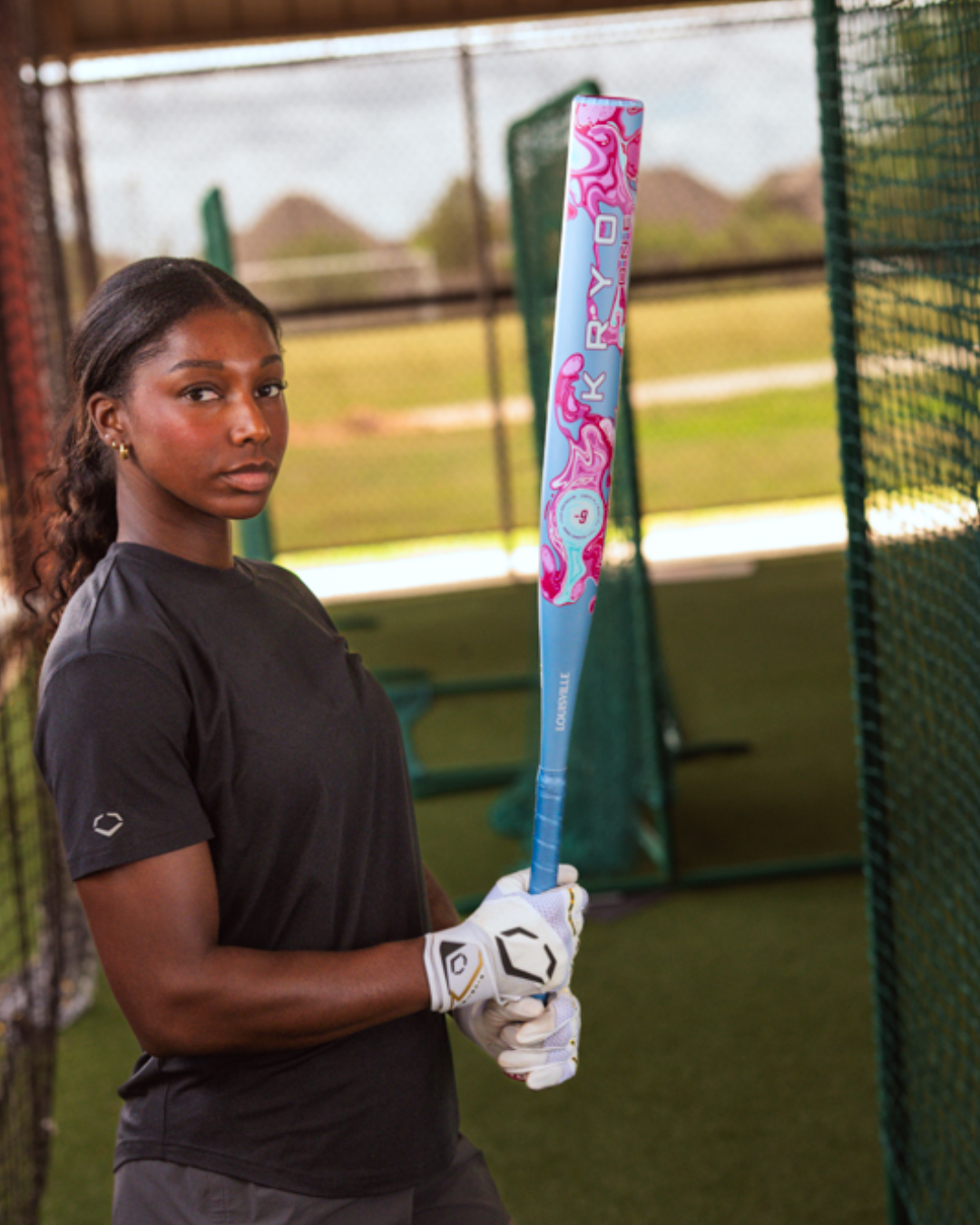 A woman wearing a black shirt and white gloves holds the Louisville Slugger Kryo One (-9) Fastpitch Softball Bat (2027, WBL4274010), standing in an outdoor batting cage.