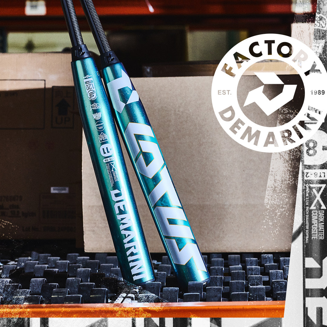 Two teal DeMarini baseball bats stand upright against cardboard boxes. A white circular Factory DeMarini Est. 1989 logo is overlaid in the top right corner. The scene has an industrial, warehouse feel.