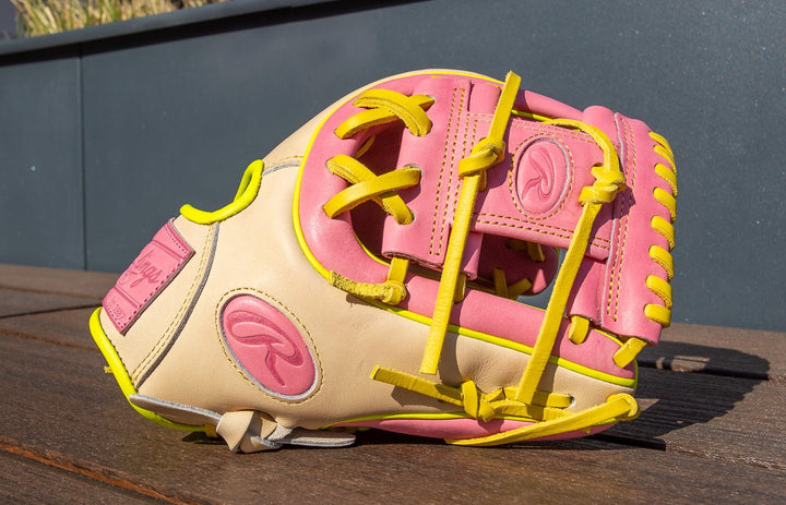 A Rawlings Heart of the Hide 11.25" Color Sync Baseball Glove (PROR312U-2P) in beige, pink, and yellow rests on a wooden surface, sunlight highlighting its vibrant Colorsync accents and fine stitching.