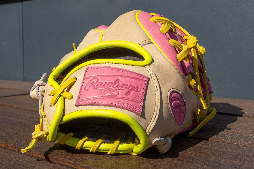 A Rawlings Heart of the Hide 11.25" Color Sync Baseball Glove (PROR312U-2P) with tan, pink, and yellow leather accents rests on a wooden surface in sunlight.