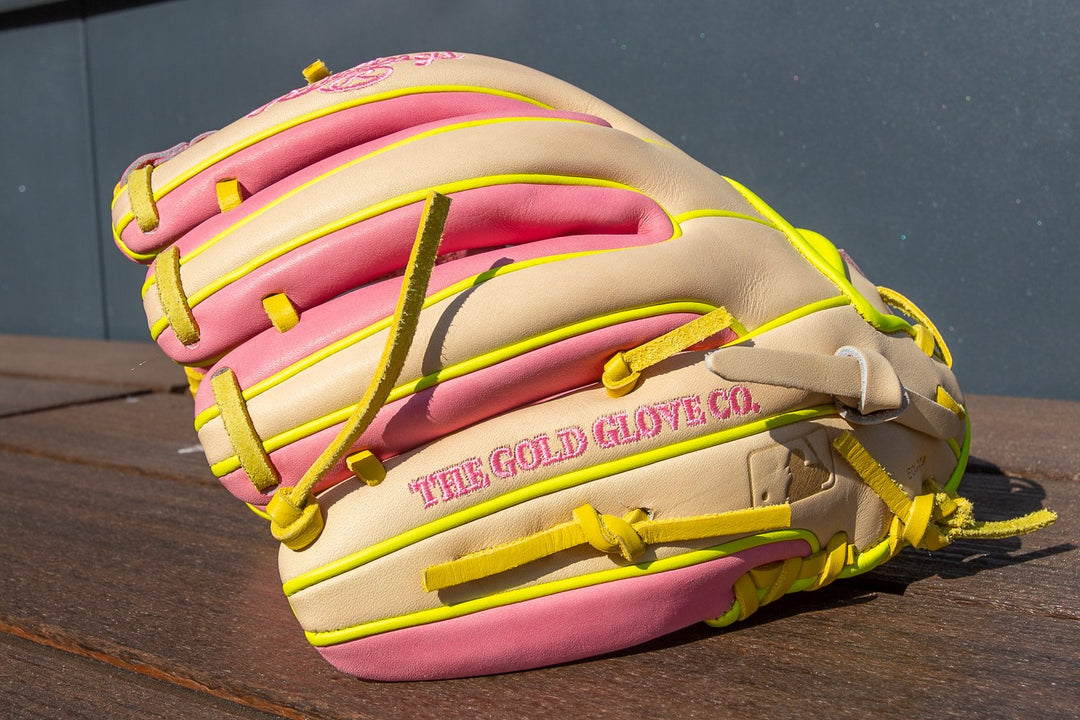 A Rawlings Heart of the Hide 11.25" Color Sync Baseball Glove (PROR312U-2P) in beige, pink, and yellow with âTHE GOLD GLOVE CO.â embroidered in pink sits on a wooden surface, its vibrant design catching the bright sunlight.