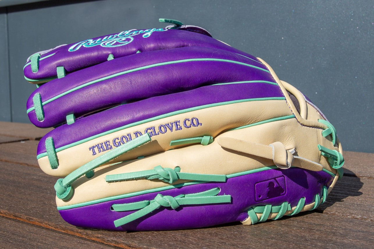 A Rawlings Heart of the Hide 12.75" Color Sync Baseball Glove (PROR3039-6CPU) in purple, cream, and teal rests on a wooden surface, featuring "THE GOLD GLOVE CO." stitched on the side.
