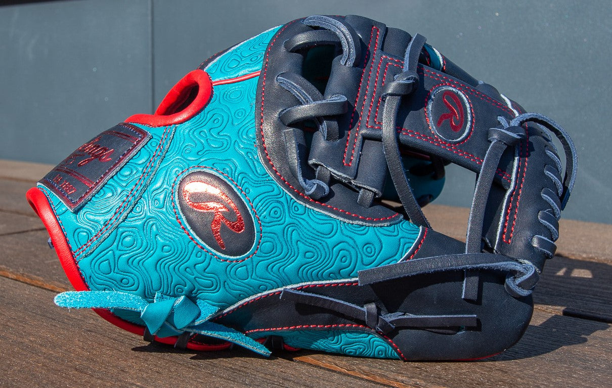 A Rawlings Heart of the Hide 11.5" Color Sync Baseball Glove (PROR204-2TE) in teal and navy with red accents rests on a wooden surface in bright sunlight.