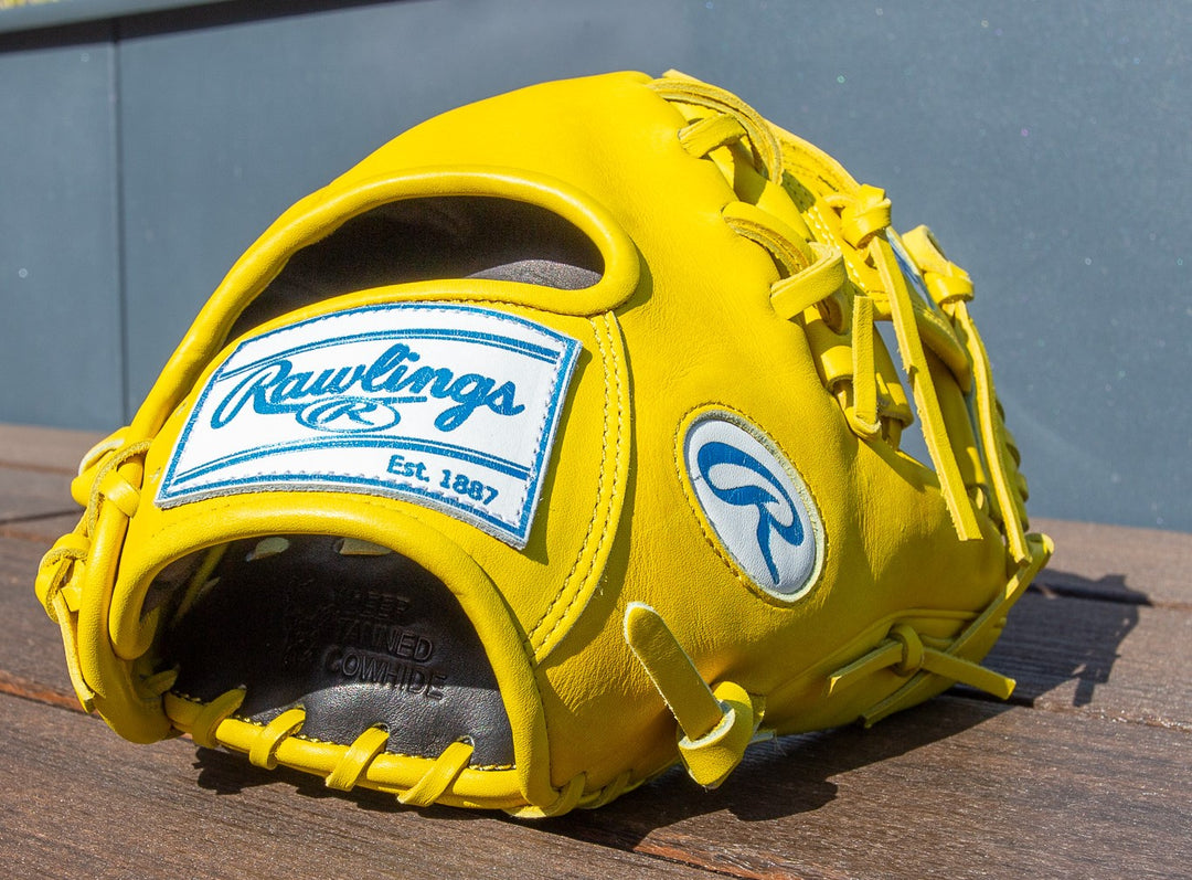 A Rawlings Heart of the Hide 11.5" Color Sync Baseball Glove (PROR204-2Y) with bright yellow leather and blue and white branding rests on a wooden surface in sunlight.