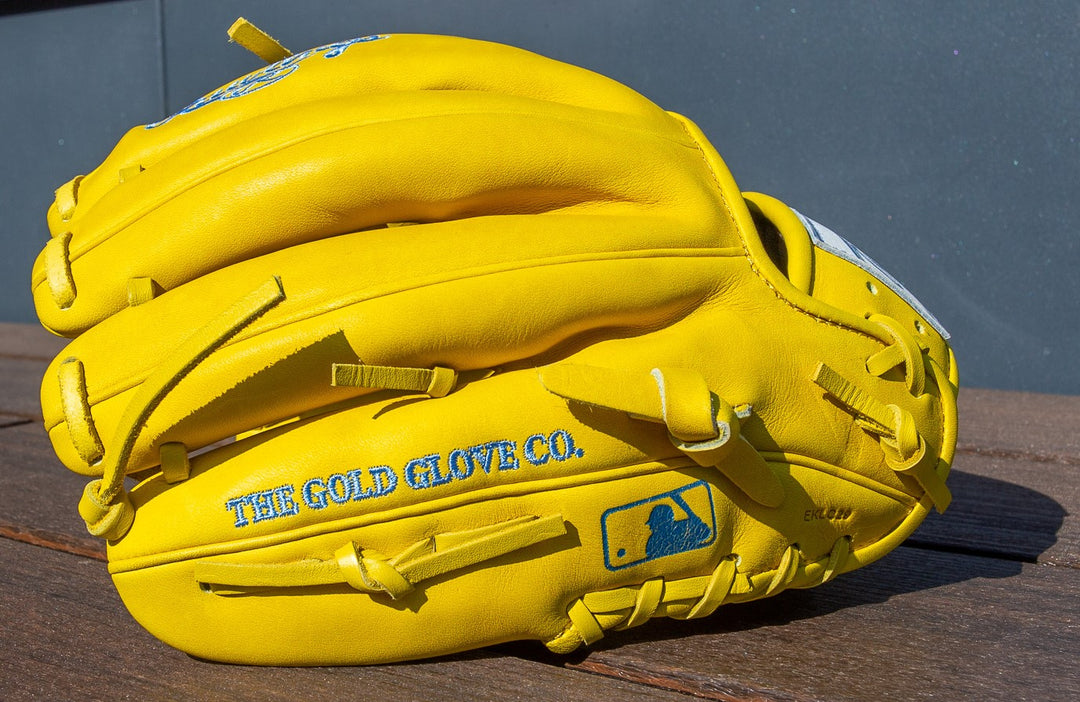 A bright yellow Rawlings Heart of the Hide 11.5" Color Sync Baseball Glove (PROR204-2Y) with THE GOLD GLOVE CO. and a blue MLB logo on the side, rests on a wooden surface against a gray background.