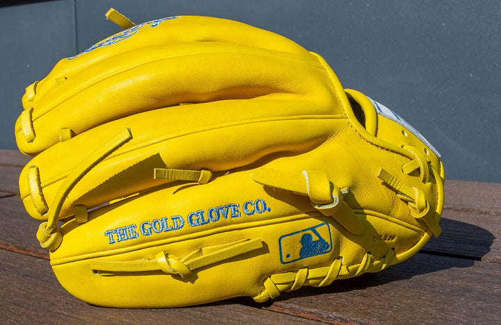 A bright yellow Rawlings Heart of the Hide 11.5" Color Sync Baseball Glove (PROR204-2Y) with THE GOLD GLOVE CO. and a blue MLB logo on the side, rests on a wooden surface against a gray background.