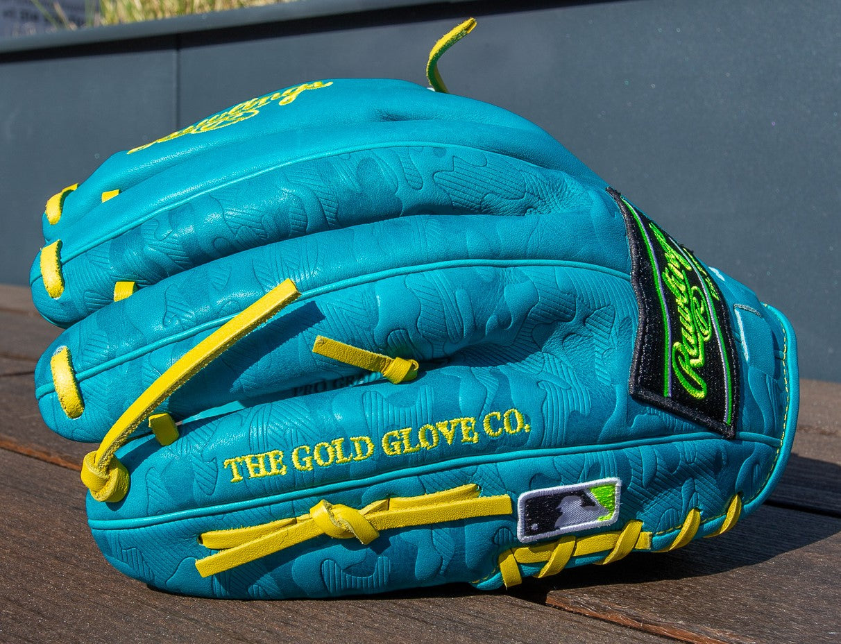 A vibrant teal Rawlings Heart of the Hide 12" Color Sync Baseball Glove (PROR206CFB-11TE) with yellow laces, embossed patterns, THE GOLD GLOVE CO., and an MLB logo in yellow rests on a wooden surface.