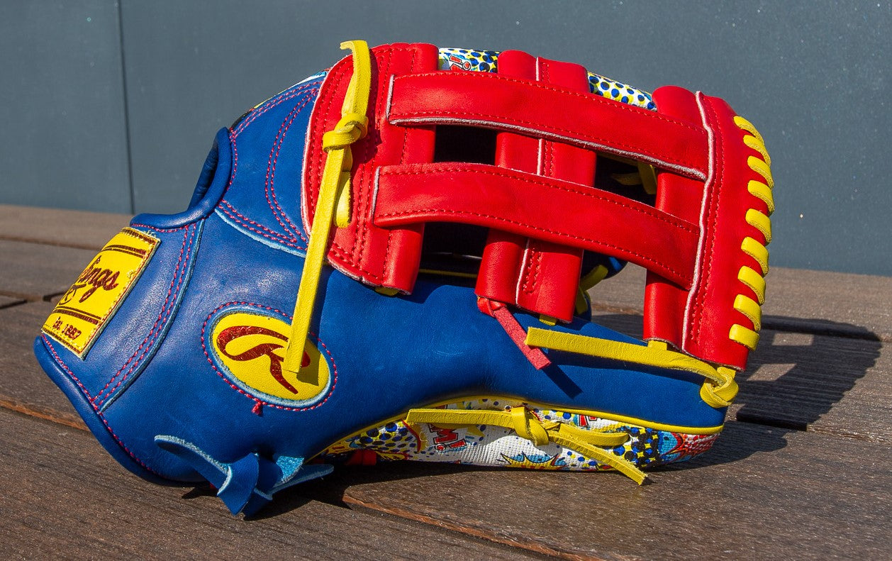 The Rawlings Heart of the Hide 12.75" Color Sync Baseball Glove (PROR3039-6RSS) features vibrant blue, red, and yellow accents with decorative patterns and bold stitching, displayed on a wooden surface against a gray background.