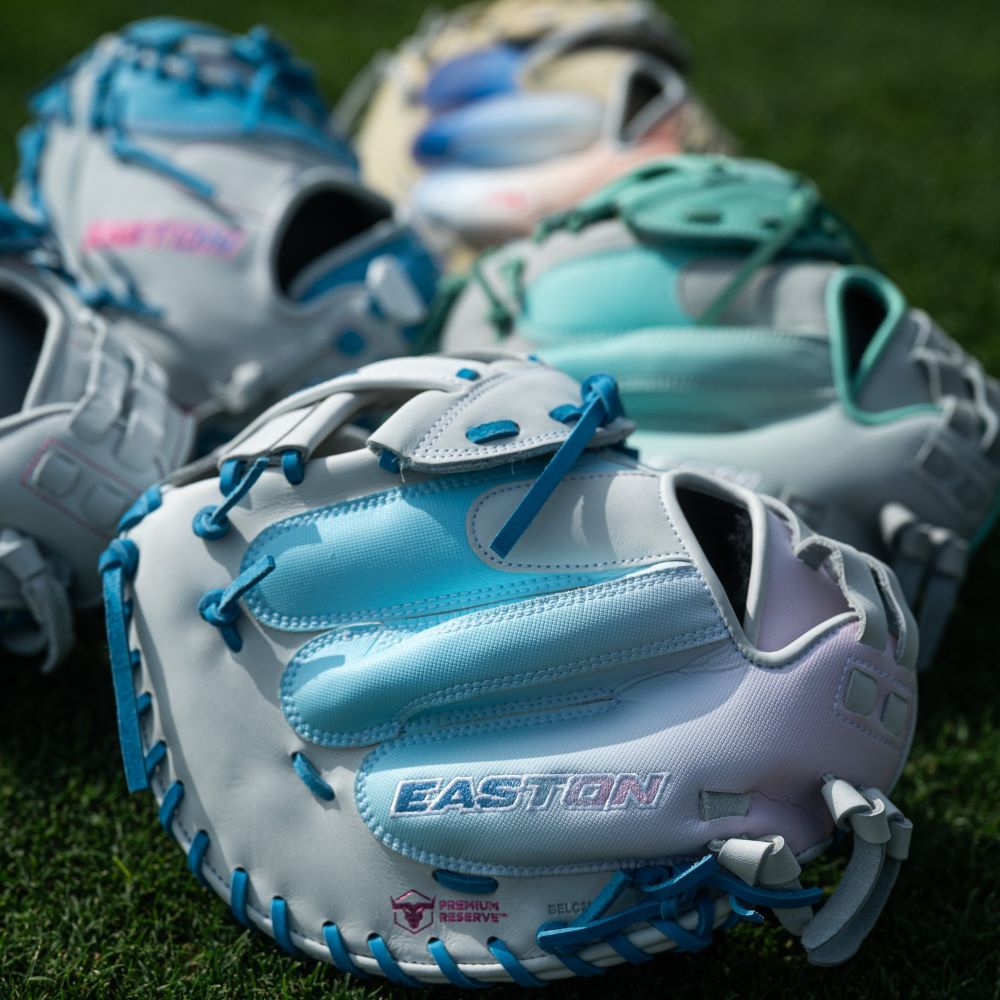 Four vibrant Easton Professional Collection Color Splash 11.75" fastpitch softball gloves (PCFP715-2WCBP) with blue, pink, and teal accents rest on green grass, the nearest displaying the Easton brand in blue and pink lettering on premium USA steerhide leather.