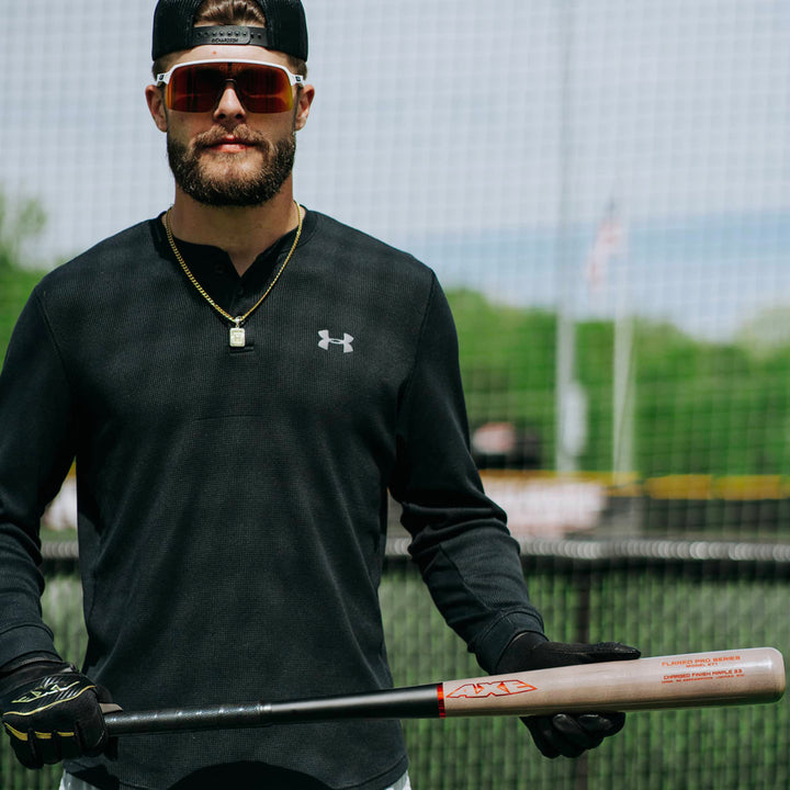 A baseball player stands on a field holding the AXE Flared Pro Series Maple Wood Baseball Bat: L124K-FLR, wearing sunglasses, a black Under Armour shirt, gloves, and a gold necklace, with a fence and trees in the background.