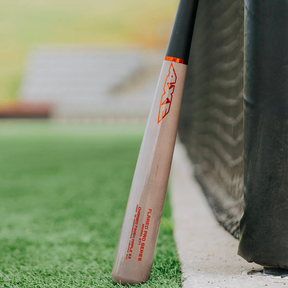 The AXE Flared Pro Series Maple Wood Baseball Bat: L124K-FLR, featuring red AXE branding and a pro flared handle, leans against a black net on a grassy baseball field with blurred stadium seating in the background.