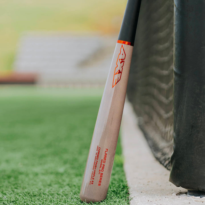 The AXE Flared Pro Series Maple Wood Baseball Bat: L124K-FLR, featuring red AXE branding and a pro flared handle, leans against a black net on a grassy baseball field with blurred stadium seating in the background.