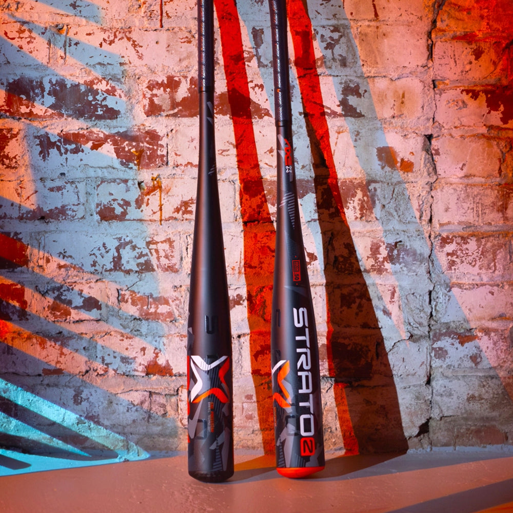 Two black baseball bats with bold red and white graphics stand upright against a textured brick wall with colorful red and blue light patterns cast across the wall and floor.