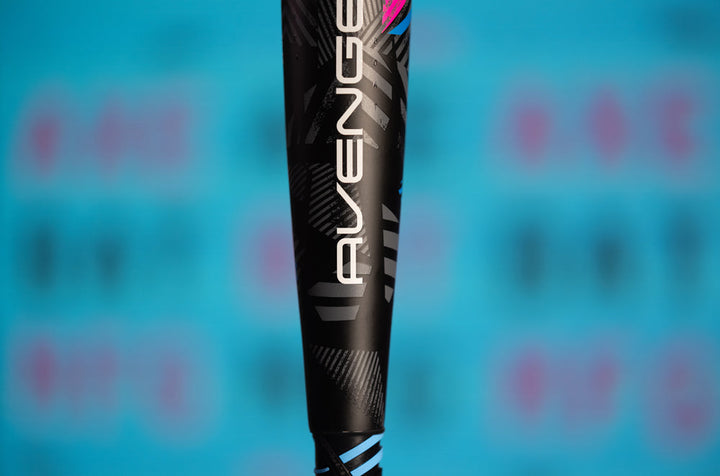 Close-up of the 2024 AXE Avenge Pro 3 Hybrid (-8) USA Baseball Bat (L174M) featuring bold white AVENGE text, set against a blue background with blurred pink and black lettering.